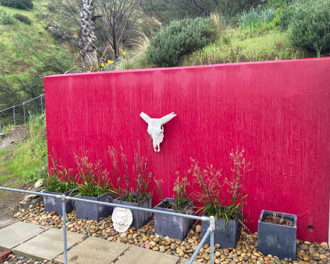 A red wall is adorned with a decorative sculpture and surrounded by planters filled with plants, set against a hillside backdrop.