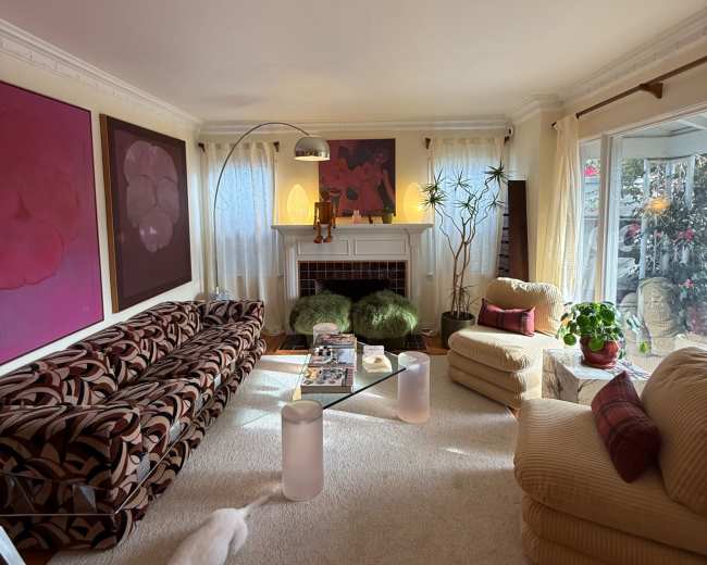 The living room features a patterned sofa, a beige armchair, a coffee table with magazines, and large windows allowing natural light to illuminate the space.