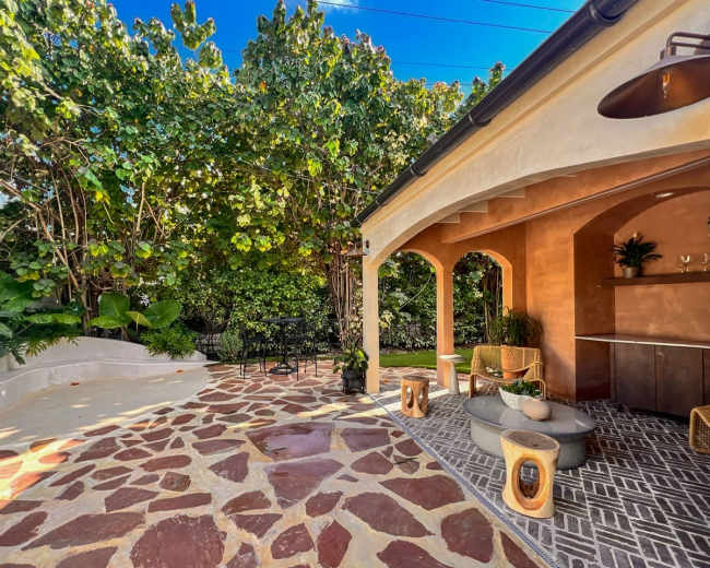 The image features a landscaped outdoor area with a stone patio, surrounded by greenery and a bar-style seating area.