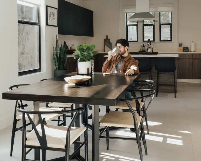 A man sits at a kitchen table, drinking from a cup, in a modern, bright dining area with plants and contemporary furnishings.