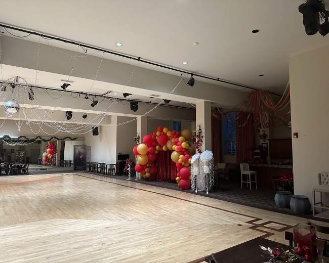 A spacious event hall decorated with red and yellow balloons, featuring a dance floor and festive decorations.