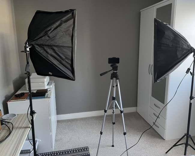 A small room with two softbox lights on stands, a tripod, and a white storage cabinet against a gray wall.