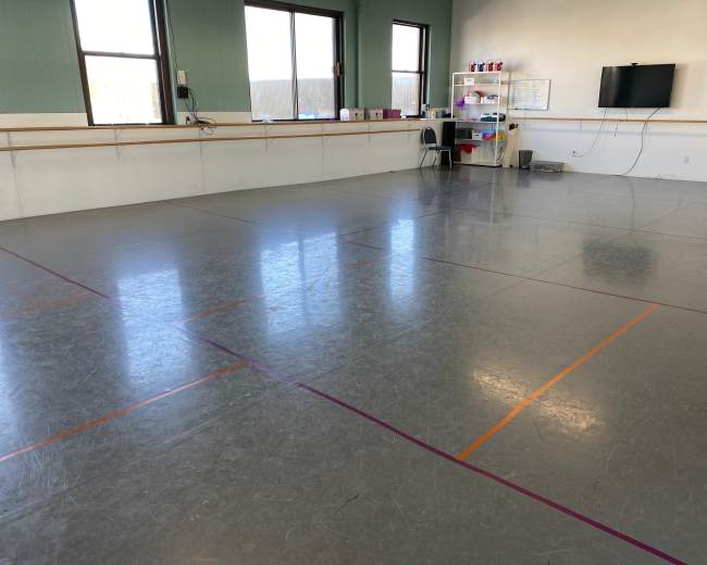 A spacious dance studio with a smooth floor marked by colored lines and a small seating area along one wall.