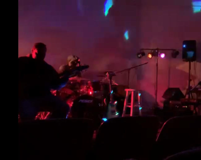 A band performs on stage while an audience watches from their seats in a dimly lit venue.