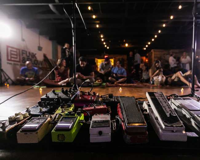 A collection of guitar effects pedals is prominently displayed in the foreground, with an audience seated in a dimly lit venue in the background.
