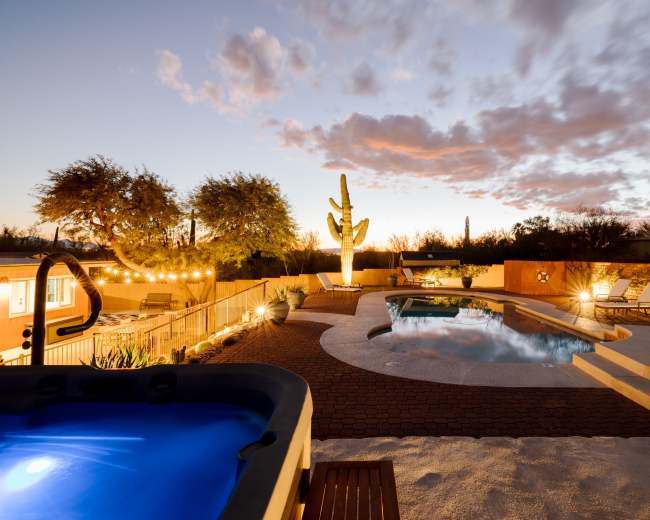 A desert backyard at dusk with a hot tub, swimming pool, and surrounding landscaping featuring a large cactus.