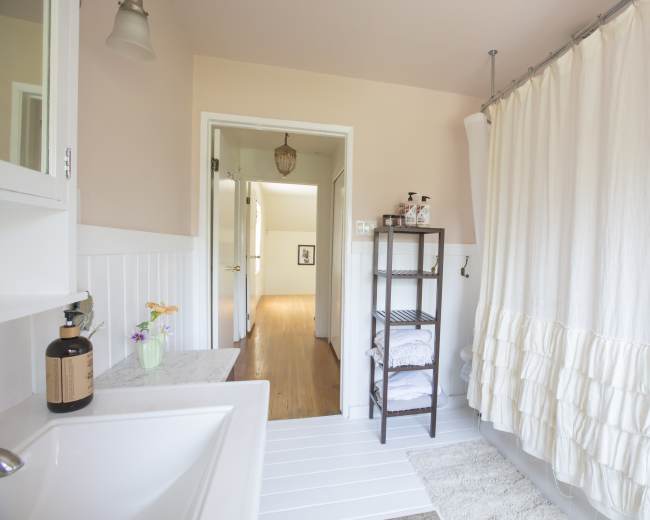 The image shows a bright bathroom with a white shower curtain, a marble countertop with a soap dispenser, and a shelf holding folded towels.