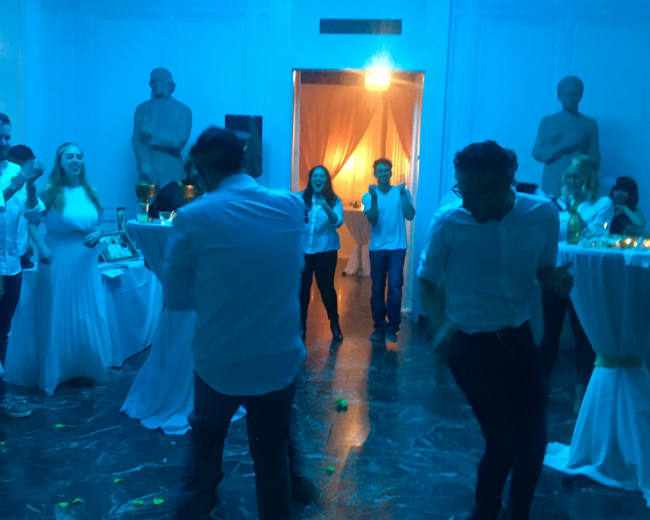 A group of people in white attire dance under blue lighting in a room with classical statues.