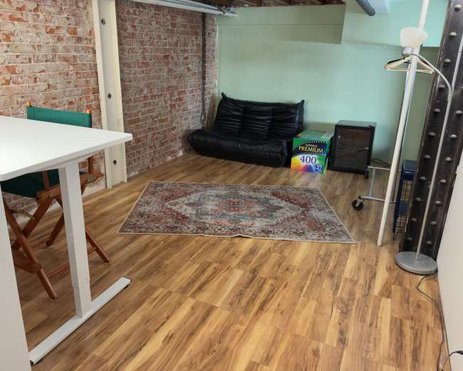 The image shows a cozy room with exposed brick walls, a wooden floor, a small table with chairs, a black bean bag chair, a rug, a floor lamp, and a storage box.