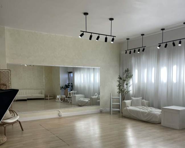 The image shows a bright, spacious studio with mirrored walls, large windows, and minimalist furniture.