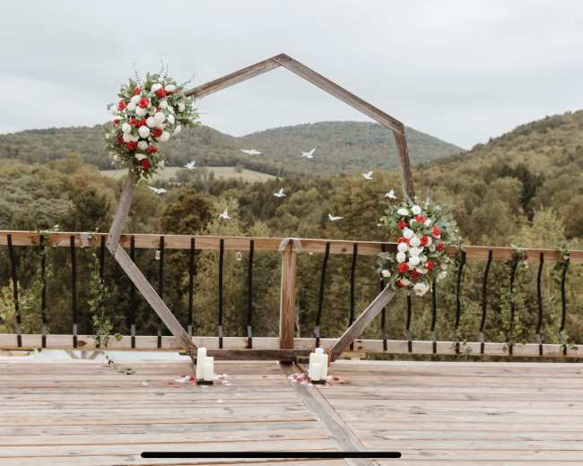 A wooden octagonal wedding arch adorned with floral arrangements and candles, set against a backdrop of green hills and a cloudy sky.