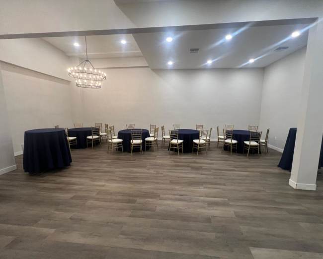 The image shows a spacious event room with several round and high-top tables covered in navy tablecloths and white chairs, illuminated by a modern chandelier.