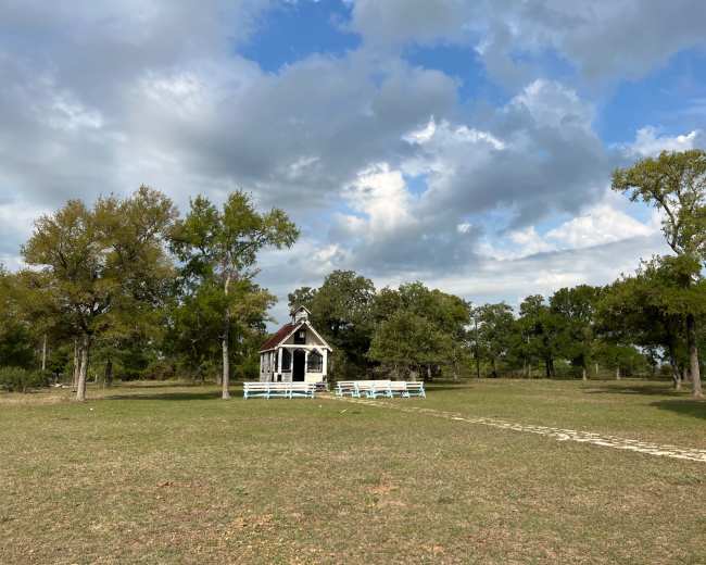 A small, rustic house with a porch stands in an open area surrounded by trees and a path made of stones leading up to it.