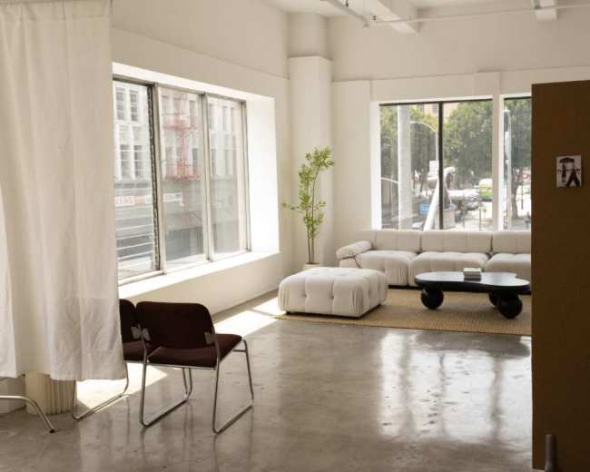 The image shows a bright, contemporary space with a white sectional couch, a round black coffee table, and large windows allowing natural light to enter.