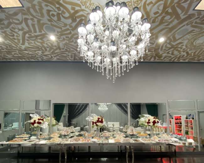 An elegantly decorated banquet hall featuring a large buffet table adorned with floral arrangements and a crystal chandelier overhead.