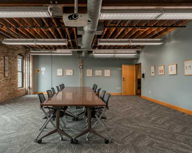 A spacious conference room featuring a long wooden table with rolling chairs, brick walls, and large windows.