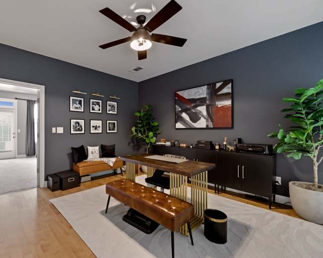 The image shows a modern office space with a dark wall, a wooden desk, a bench, and various framed artworks.
