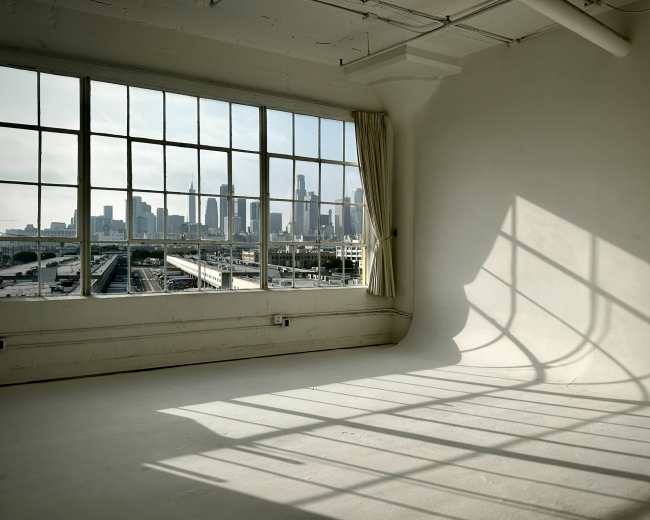 A large window offers a view of a city skyline, casting shadows onto the empty floor of a sunlit room.