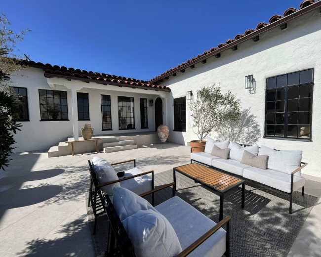 A patio with modern seating and a table is surrounded by white walls and large windows in a sunny courtyard.