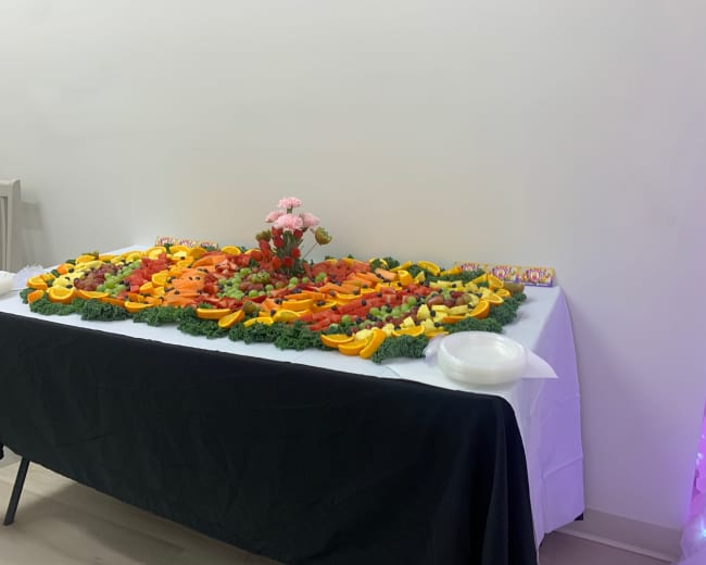 A table is filled with an array of colorful fruit arranged in an artistic pattern, surrounded by decorative greens and featuring a small bouquet of flowers.