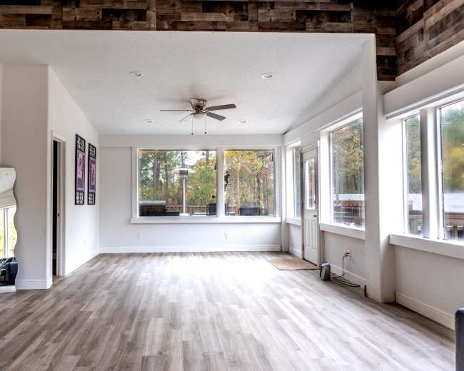 The image shows a spacious, bright interior room with large windows, a ceiling fan, and light-colored wooden flooring.