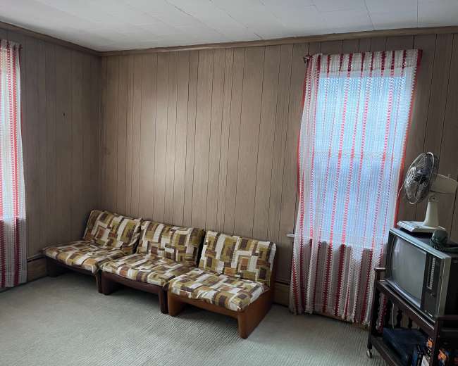 The image shows a small living room with a patterned sofa, two windows covered in striped curtains, and a television set on a stand.