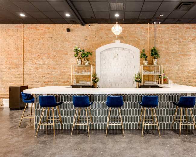 The image shows a modern bar with a white countertop, blue upholstered stools, and a textured stone wall in the background.