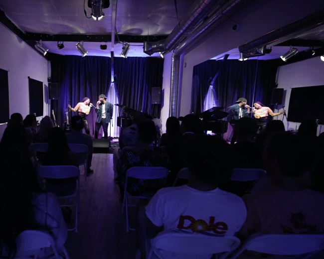 A performance is taking place on a stage with blue curtains, while an audience sits in white chairs facing the musicians.