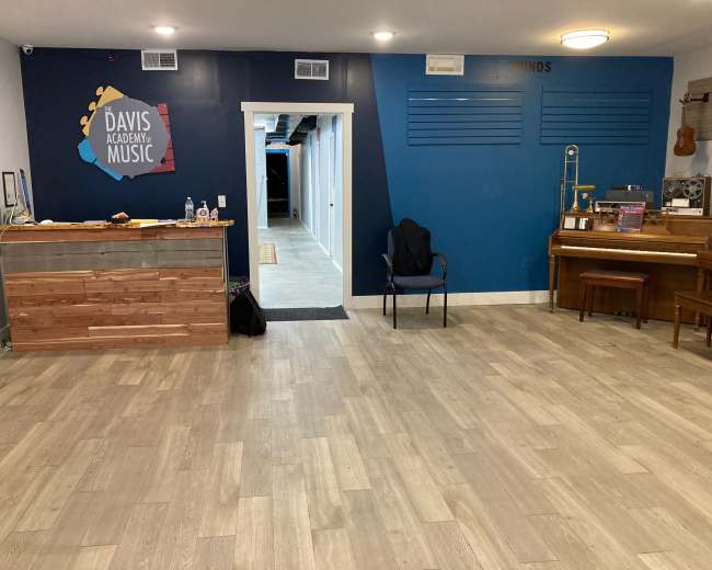 The image shows a music academy reception area with a wooden counter, a chair, a piano, and a blue accent wall featuring the academy's logo.