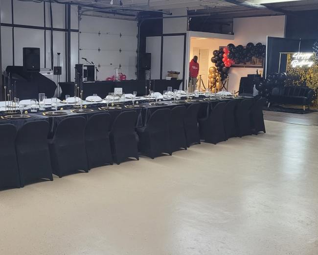 A long dining table set for a formal event is positioned in a spacious venue with black tablecloths and decorations.