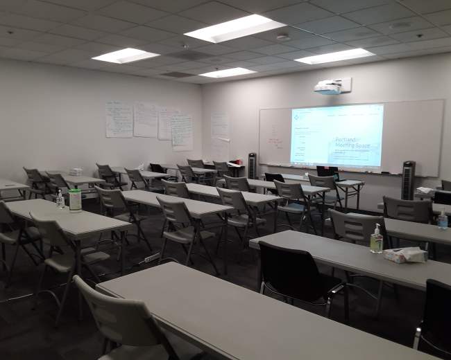 A meeting room is set up with rows of tables and chairs, featuring a projector screen displaying information about a Portland meeting space.