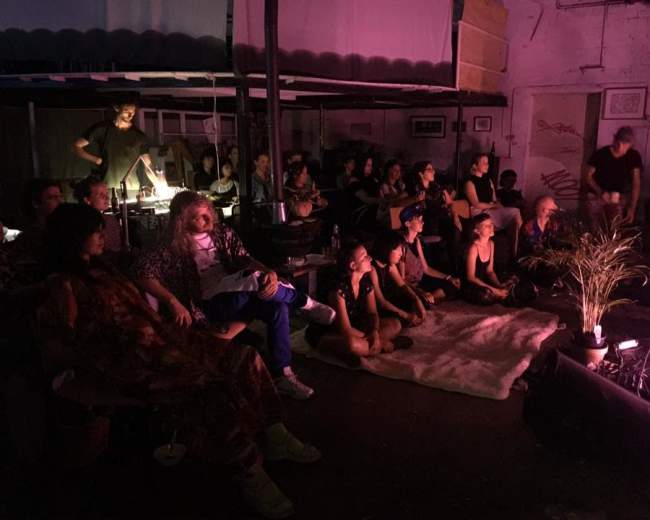 A group of people is seated in a dimly lit room, some facing a performance area while others appear engaged in conversation.