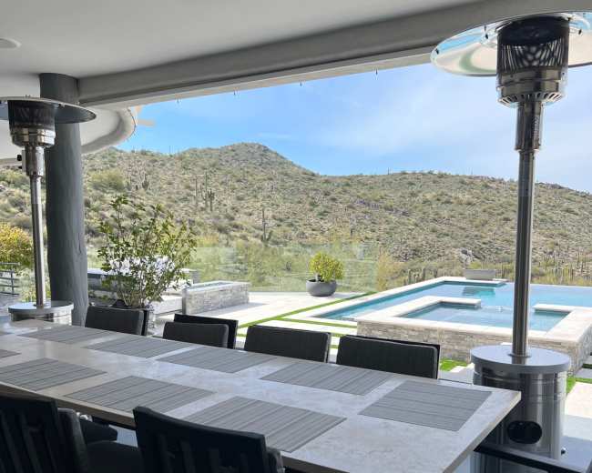 A dining table is set on a patio overlooking a pool and mountainous desert landscape.