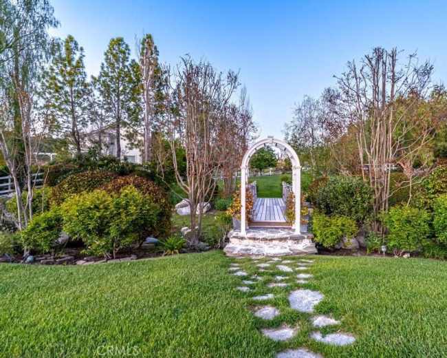 A stone pathway leads to a white archway surrounded by greenery and trees in a garden setting.