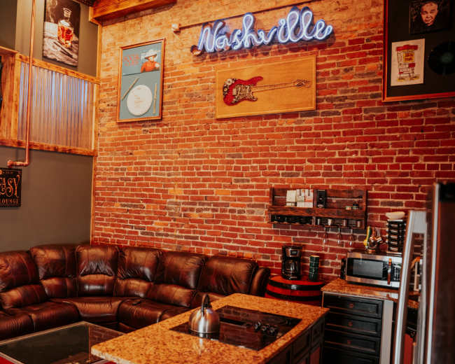 The interior features a cozy seating area with a dark leather couch, a granite coffee table, and a wall adorned with exposed brick and various musical decorations, including a neon sign that says "Nashville."