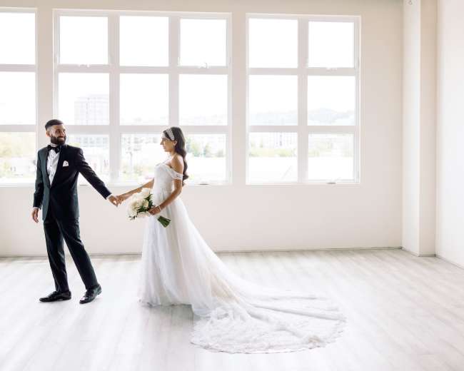 A bride and groom hold hands and walk together in a spacious, well-lit room with large windows.