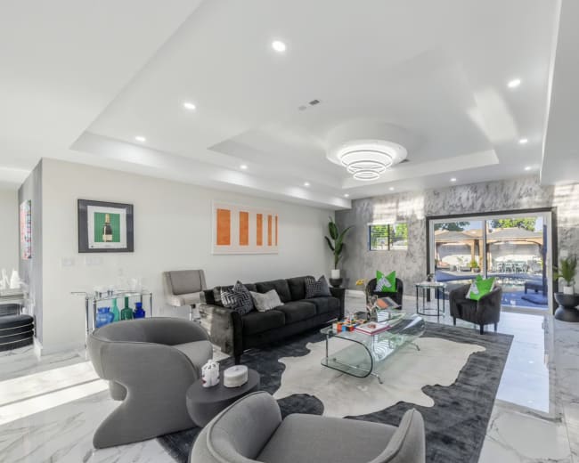 The living room features modern furniture, a glass coffee table, and large windows that open to an outdoor patio.
