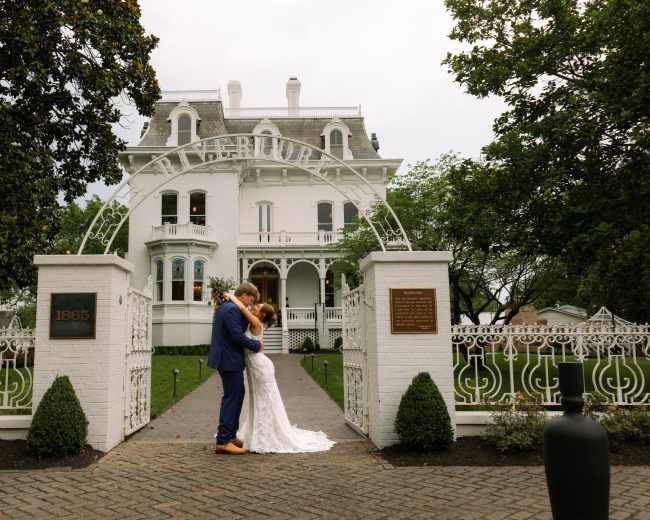 A newlywed couple embraces in front of a large white Victorian house with an ornate archway and grassy lawn.