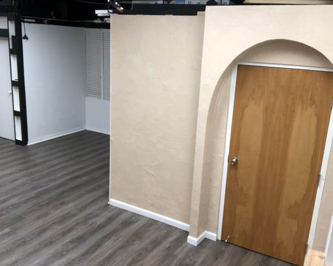 The image shows a spacious interior room with light-colored walls, a wooden door with an archway, and a wooden floor.