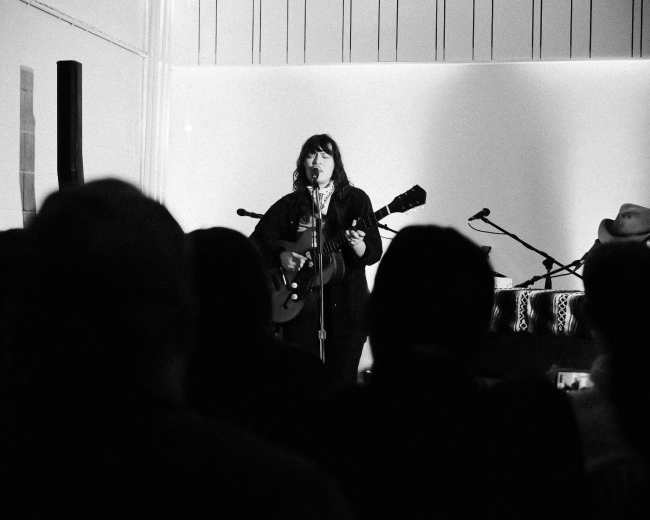 A musician stands on stage, playing a guitar while an audience watches from the foreground.