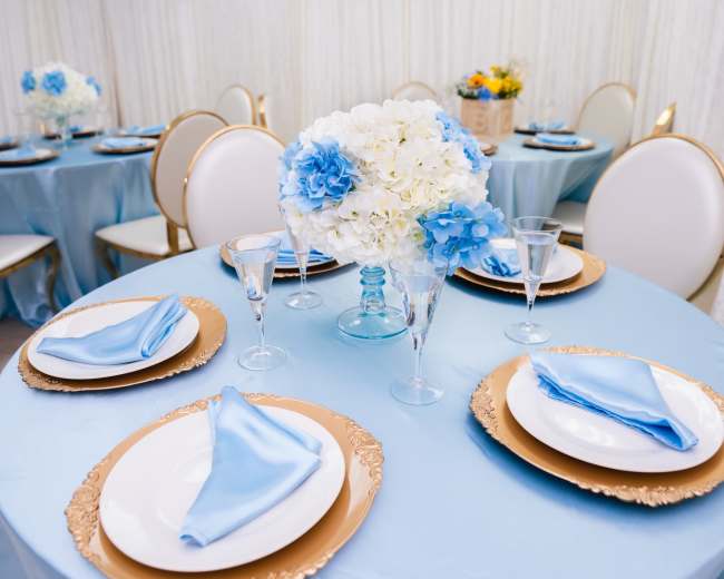 A neatly arranged banquet setup features round tables with blue tablecloths, white and blue floral centerpieces, and coordinated plates and napkins.