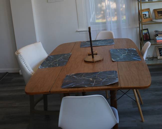 A wooden dining table with four chairs is set with placemats underneath a decorative light fixture and a nearby shelf holds framed photos.