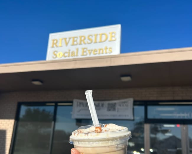 A person holds a cup of iced coffee in front of a building with a sign that reads "RIVERSIDE Social Events" under a clear blue sky.