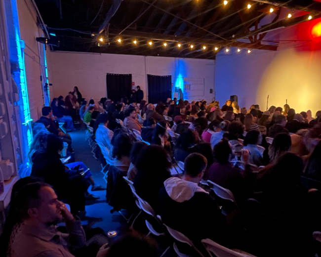 A large audience gathers in a well-lit room decorated with string lights, facing a stage set for an event.
