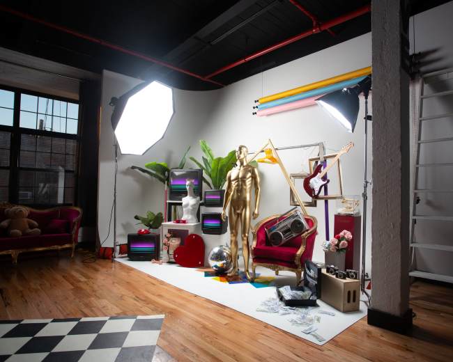 The image shows a creatively designed studio space featuring a golden mannequin, colorful decor, various musical instruments, and large light sources.