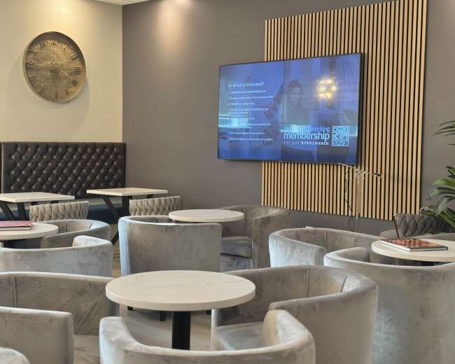 The image shows a modern lounge area with grey velvet seating, small white tables, and a large screen displaying information on a wall.