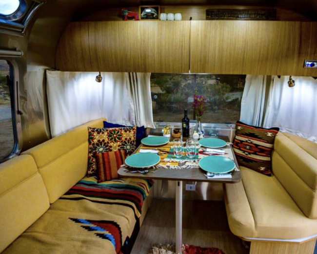 The image shows a cozy dining area inside a retro-style trailer, featuring a table set for a meal with colorful plates and decorative cushions.