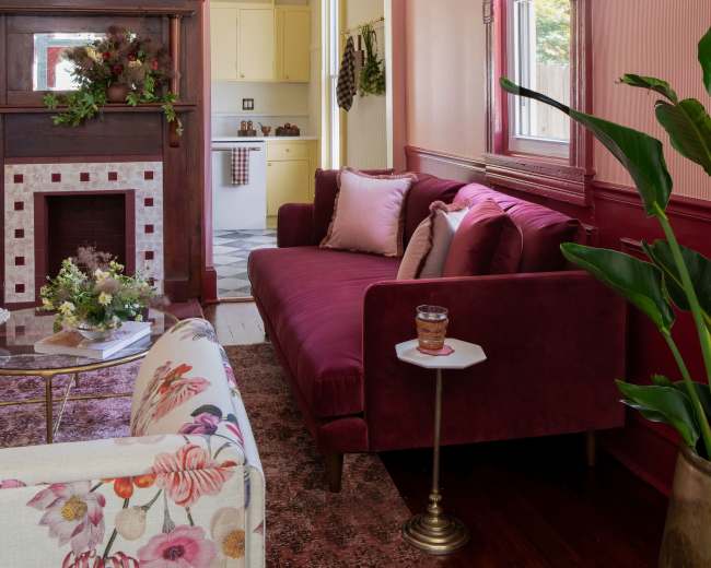 The image shows a cozy living room with a burgundy couch, a floral armchair, and a decorative fireplace, leading to a yellow kitchen visible in the background.