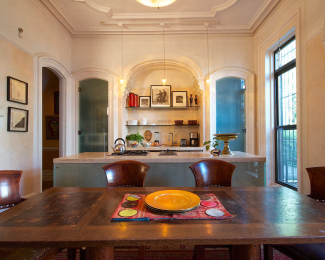 The image shows a dining area with a wooden table set with a yellow plate, surrounded by four chairs, and a kitchen area visible in the background.