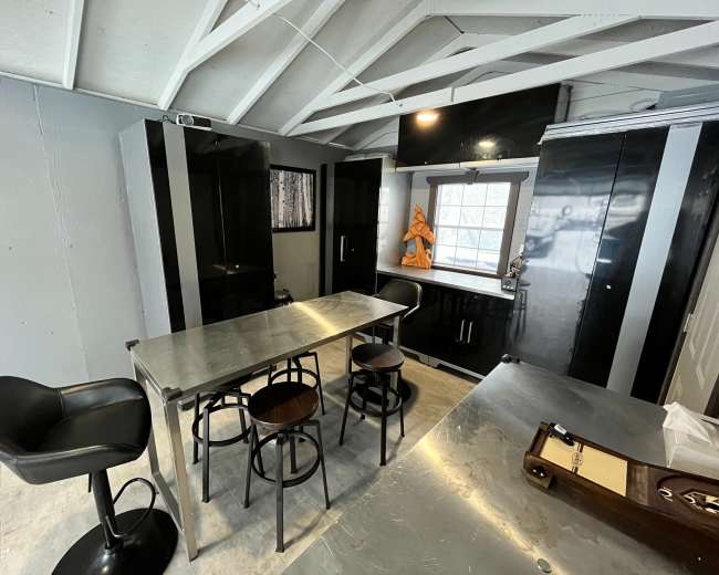 A modern kitchen features sleek black cabinets, a metallic table, and bar stools under a slanted ceiling with exposed beams.